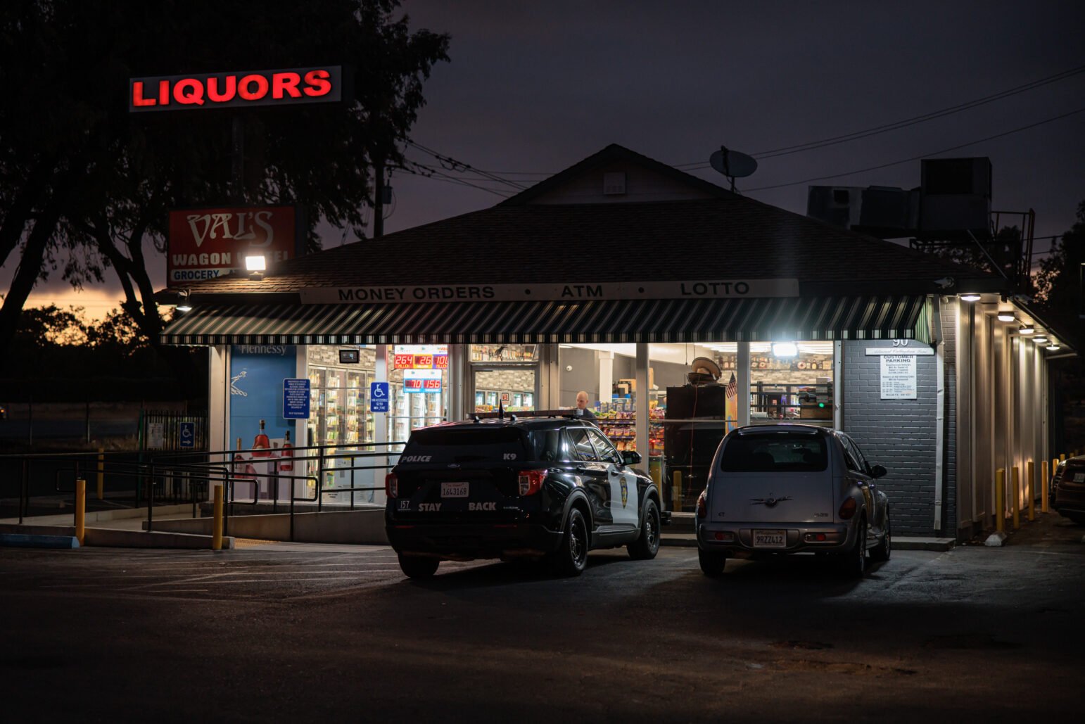 A Vallejo police SUV and a silver PT Cruiser are parked outside Val’s Wagon Wheel Liquors at dusk. The store’s bright interior contrasts with the darkened sky, and a red neon “LIQUORS” sign glows above the building.