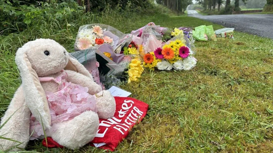 A teddy bear bunny, sat on top of a red Arsenal jersey. They are alongside a bunch of flowers. All have been placed on the grass on the side of a road.